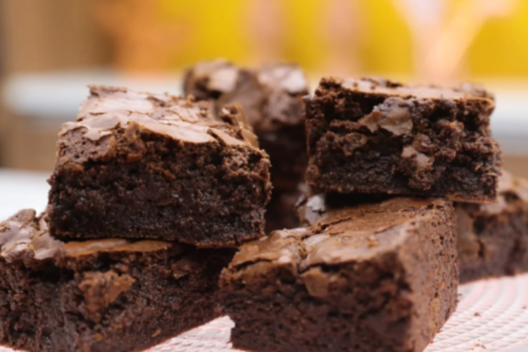 Brownies caseiros cortados em pedaços quadrados, com crosta brilhante e textura úmida no centro, servidos em um prato rústico com xícara de café ao fundo.