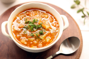 "Prato fundo com sopa de legumes caseira, contendo pedaços de cenoura, batata, ervilha e vagem, decorada com salsinha, ao lado de pão integral e ingredientes frescos sobre uma mesa de madeira."